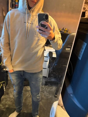 Beige hoodie från Calvin Klein Jeans - Säljer en beige hoodie från Calvin Klein Jeans med klassisk huva och dragsnören. Diskret CK-logga broderad på bröstet. Hoodien har ribbade muddar vid ärmslut och nederkant. Perfekt för en avslappnad och stilren look.