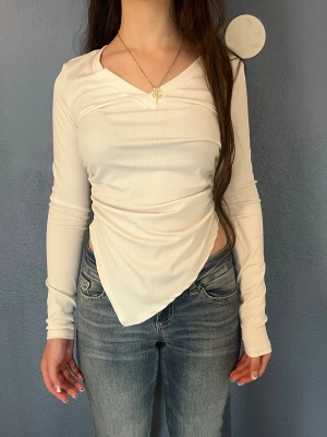 Vit ribbad omlottopp med lång ärm - Trendig vit ribbad topp med omlottdetalj och asymmetrisk nederkant. Långärmad och croppad modell som ger en snygg siluett. Perfekt att styla med jeans eller kostymbyxor för en cool look.