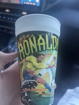 Plastmugg med Ronaldinho-motiv - Cool plastmugg med färgglad tecknad bild av en fotbollsspelare i gul tröja och blå shorts samt texten 'RONALDI'. Perfekt för dig som gillar fotboll och vill ha något unikt till drycken. Muggen har vit kant upptill och är dekorerad med stjärnor och fotbollstema.
