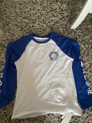 Blåvit långärmad topp Chrome Hearts - Snygg långärmad topp från Chrome Hearts med blå ärmar och vit front. Tryck på bröstet och längs ärmarna. Klassisk baseball-inspirerad design med rund halsringning. Perfekt för dig som gillar streetwear och vill sticka ut.