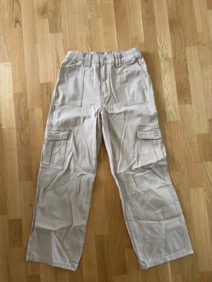 Beiga cargobyxor med fickor - Snygga beiga cargobyxor med raka ben och flera praktiska fickor på sidorna och baktill. Byxorna har elastisk midja och är tillverkade i ett mjukt bomullstyg. Perfekta för en avslappnad och trendig stil.