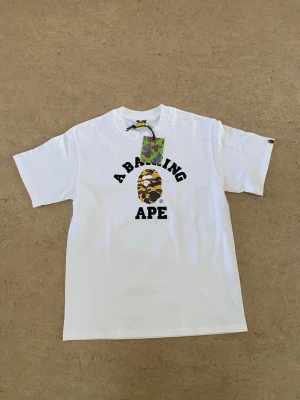 Vit BAPE t-shirt med camo-logga - Snygg vit t-shirt från A Bathing Ape med klassisk camo-logga och texten 'A BATHING APE' på bröstet. T-shirten har rund hals, korta ärmar och är tillverkad i mjuk bomull. Perfekt för dig som gillar streetwear och vill sticka ut med en ikonisk design. TAGS OCH PÅSE FÖLJER MED!‼️