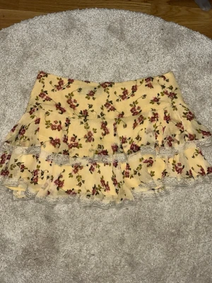 Blommig volangkjol från Zara - Supersöt kort kjol från Zara i ljusgul färg med blommigt mönster i rött och grönt. Kjolen har dubbla volanger och spetsdetaljer längs kanterna. Perfekt för vår och sommar, och ger en romantisk vibe till din outfit.