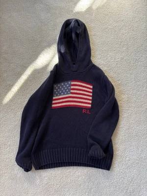 Mörkblå hoodie från Ralph Lauren - Mörkblå stickad hoodie från Ralph Lauren med stor amerikansk flagga på bröstet och RL-broderi under. Tröjan har huva och ribbade muddar. Perfekt för dig som gillar klassisk och preppy stil.