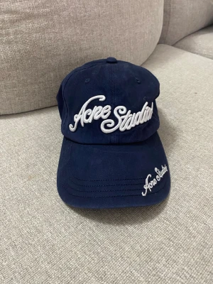 Mörkblå keps från Acne Studios - Snygg mörkblå keps från Acne Studios med vit broderad logga framtill och på skärmen. Klassisk böjd skärm och justerbart spänne bak. Tillverkad i bomullstyg som ger en avslappnad och trendig look.