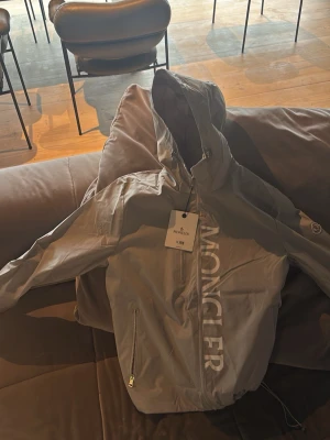 Grå vindjacka från Moncler - Snygg grå vindjacka från Moncler med huva och dragkedja framtill. Jackan har Moncler-logga på ärmen och stort tryck med märkesnamnet längs framsidan. Perfekt för blåsiga dagar och streetwear-stil.