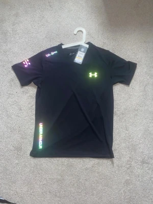 Svart Under Armour t-shirt - Snygg svart t-shirt från Under Armour med korta ärmar och reflekterande logga och textdetaljer i neonfärger. Storlek M men kan passa både M och S