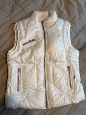 Trendig vit pufferväst från Vero Moda - Denna klassiska pufferdesign har en hög krage för extra värme och en dragkedja framtill.  Flera fickor med dragkedja ger gott om förvaring, och den mjuka och sköna insidan garanterar komfort. Snygga sömmar ger en fin passform.  Skick: Mycket fint skick – varsamt använd, inga synliga defekter.  Storlek L, men passar som M för en mer perfekt/normal passform.  Modellen säljs inte längre – svår att få tag på!