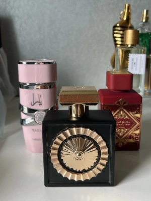 Paketpris parfymer  - Säljer en exklusiv parfym i en fyrkantig svart flaska med detaljer i guld. Parfymen är inspirerad av Billie Eilish, Eilish. Den är 100ml. Flaskan har ett unikt, dekorativt mönster framtill och ett elegant guldfärgat lock. Perfekt för dig som gillar snygg design och vill ha något som sticker ut på parfymhyllan. Max 30% av parfymen är använd, alltså är den nästan full. På köpet tillkommer även 2 andra använda parfymer från Lattafa. Minst 50% av innehållet kvarstår i de. De ingår. 