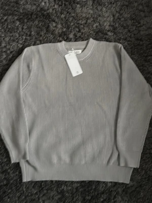 Ljusgrå stickad tröja maison margiela  - En stilren ljusgrå stickad tröja med rund hals och ribbade muddar. Tröjan har en enkel och clean design som passar till det mesta. Perfekt för lager på lager och funkar året runt. Materialet ser ut att vara mjukt och bekvämt.