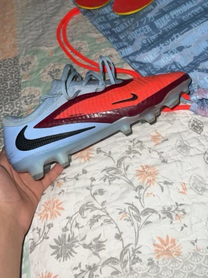 Nike fotbollssko röd och ljusblå - Säljer en Nike fotbollssko med snygg design i rött, vinrött och ljusblått. Skon har svarta Nike-loggor på sidan och är tillverkad i syntetmaterial med snörning och dobbar för spel på gräs. Perfekt för dig som vill sticka ut på planen. De är elite också 