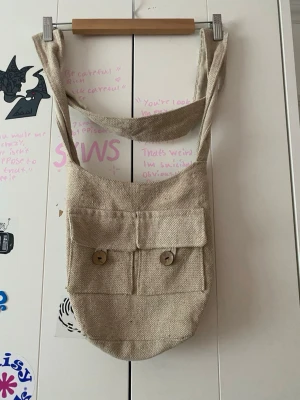 Beige shoppingväska i linne - Säljer en beige shoppingväska i grovt linnetyg med bred axelrem och två stora fickor framtill som stängs med träknappar. Perfekt för dig som gillar en enkel och naturlig stil. Väskan har en avslappnad form och rymmer mycket.