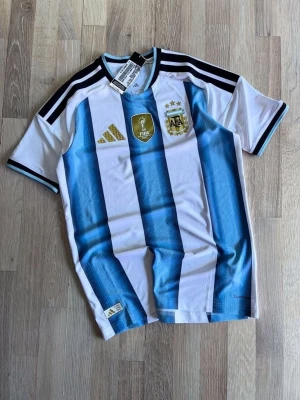 Argentina matchtröja Adidas herr - Snygg Argentina matchtröja från Adidas med klassiska blå och vita ränder, FIFA-märke och AFA-logga på bröstet. Tröjan har svarta detaljer på axlarna och korta ärmar. Perfekt för fotbollsfans som vill visa sitt stöd för landslaget.
