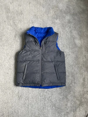 Grå dunväst från Polo Ralph Lauren - Snygg grå dunväst från Polo Ralph Lauren med blå insida och hög krage. Västen har två fickor med dragkedja och diskret broderad logga på bröstet. Perfekt för lager-på-lager och håller dig varm under kyliga dagar.