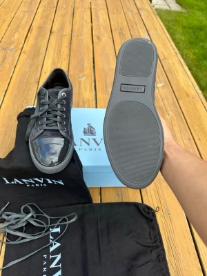 Svarta Lanvin sneakers med lackdetalj - Snygga svarta sneakers från Lanvin Paris med glansig lackdetalj på tån och klassisk snörning. Skorna har en låg profil och är tillverkade i en mix av mocka och läder. Kommer med originalpåse och extra skosnören. Perfekta för dig som gillar exklusiv streetstyle.