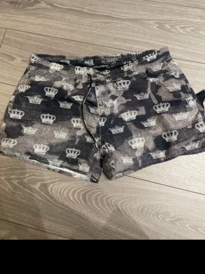 Camouflage shorts från Dolce & Gabbana - Snygga shorts från Dolce & Gabbana med grått och svart camouflage-mönster och vita kronor över hela tyget. Shortsen har snörning i midjan och är perfekta för dig som vill sticka ut med en unik design.