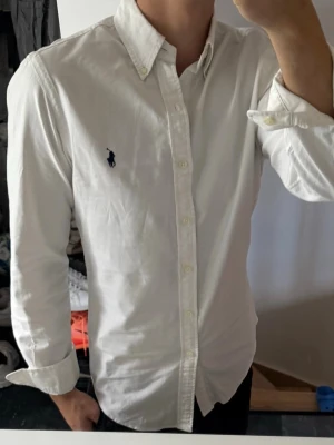 Vit skjorta från Ralph Lauren - Klassisk vit skjorta från Ralph Lauren med button-down krage och broderad logga på bröstet. Skjortan har långa ärmar, knappar framtill och är tillverkad i bomull för en skön och stilren look. Lite missfärgad annars bra skick 