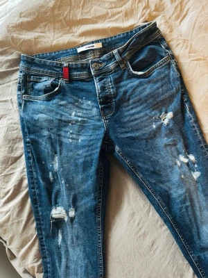Blå Distress Jeans | 2YPremium - Säljer ett par blå jeans från 2YPremium med snygga slitningar och färgstänk för en trendig look. Jeansen har klassisk tight model, 32 midja och 30 längd. Perfekta för dig som vill stå ut i sommar.