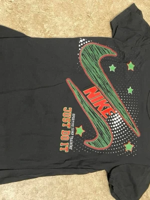 Svart Nike t-shirt med grafiskt tryck - Svart t-shirt från Nike med stort grafiskt tryck i grönt, rött och vitt på framsidan. Trycket har Nike-loggan, texten 'Just Do It' och stjärnor i grönt och rött. Klassisk rund hals och korta ärmar. Perfekt för dig som gillar sportig streetstyle.