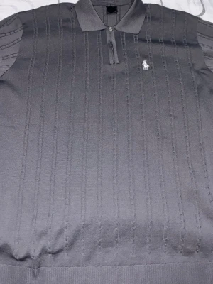 Grå polotopp från Polo Ralph Lauren - Snygg grå polotopp från Polo Ralph Lauren med diskret vertikalt mönster och klassisk krage. Toppen har korta ärmar, half zip-dragkedja och den ikoniska loggan broderad på bröstet. Perfekt för en stilren och avslappnad look.