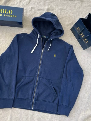 Marinblå hoodie från Polo Ralph Lauren - Snygg marinblå hoodie från Polo Ralph Lauren med dragkedja, vit dragsko i huvan och klassisk gul logga på bröstet. Hoodien har känguruficka framtill och är tillverkad i mjukt bomullsmaterial. Perfekt för en avslappnad och stilren look.