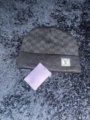 Svart mössa med mönster  - Snygg svart mössa från L. V med diskret rutmönster och vikt kant. Märkt med L. V-logga framtill och etikett på insidan. Tillverkad i mjukt stickat material som känns lyxigt och stilrent. Perfekt accessoar för dig som gillar exklusiva detaljer.