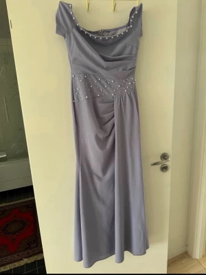 Ljuslila långklänning med pärlor - Elegant ljuslila långklänning med pärldetaljer längs urringningen och midjan. Klänningen har offshoulder-modell och draperad överdel som ger en snygg siluett. Perfekt för bal eller festliga tillfällen. Storlek M