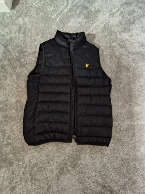 Svart dunväst från Lyle & Scott - Snygg svart dunväst från Lyle & Scott med gul broderad logga på bröstet. Västen har hög krage, dragkedja framtill och quiltad design som ger en sportig look. Perfekt att slänga över en hoodie eller tröja för extra värme.