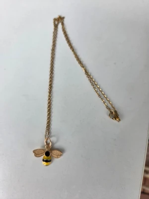 Guldigt halsband med bi - Halsband i guldfärgad metall med ett hänge format som en bi. Kedjan är reglerbar upp till 45 cm. 