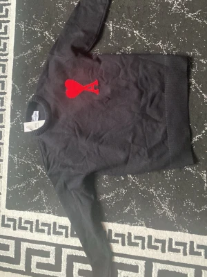 Svart stickad tröja från Ami Paris - Snygg svart stickad tröja från Ami Paris med röd hjärtlogga och bokstaven A på bröstet. Tröjan har rund halsringning och ribbade muddar vid ärmslut och nederkant. Perfekt för dig som gillar stilrena plagg med en twist. Storlek M men passar S 