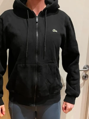Svart zip hoodie från Lacoste - Säljer en svart hoodie från Lacoste med dragkedja framtill och klassisk krokodil-logga på bröstet. Hoodien har huva med dragsko, två stora fickor fram och ribbade muddar vid ärmar och midja. Tillverkad i mjukt bomullsmaterial för skön känsla.