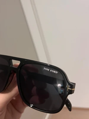 Svarta solglasögon från Tom Ford - Snygga svarta solglasögon från Tom Ford med klassisk fyrkantig form och mörka glas. Bågarna har gulddetaljer vid gångjärnen och loggan på linsen. Perfekta för dig som vill ha en stilren och exklusiv look.