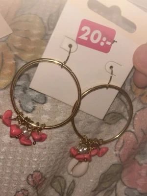 Guldiga hoops med rosa stenar och snäcka - Stora guldiga hoop-örhängen med hängande rosa stenar och små pärlor. Ena örhänget har även en vit snäcka som detalj. Perfekta för dig som gillar boho-stil och färgglada accessoarer.