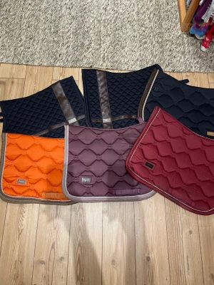 Färgglada schabrak till häst - Säljer ett gäng schabrak i olika färger: svart, orange, vinröd och brun. Alla har quiltad design och kantband i kontrastfärg. Perfekt för dig som vill variera stilen på din hästutrustning. Några har detaljer i konstläder. Är från equstrian Stockholm.