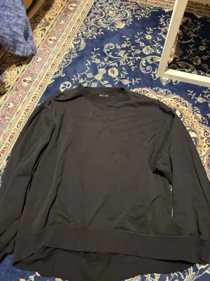 Svart sweatshirt - En enkel svart sweatshirt med rund hals och långa ärmar. Tröjan har en avslappnad passform och ribbade muddar vid ärmslut och nederkant. Perfekt för en casual och stilren look. Materialet känns mjukt och bekvämt mot huden.