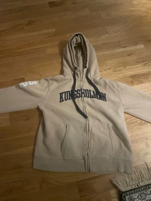 Beige hoodie med tryck KUNGSHOLMEN - Beige ziphoodie med mörkblå texten 'KUNGSHOLMEN' på bröstet och siffran 29 på ärmen. Hoodien har dragkedja, två fickor fram och justerbar huva med mörka snören. Mjuk insida i bomullsmix, perfekt för chill dagar.