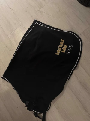 Svart fleece länd täcke med SWE och kronor - Svart fleecetäcke med broderade tre kronor och texten SWE i vitt. Täcket har vita kantband och är format för att passa häst. Perfekt för dig som vill visa svensk stil i stallet.