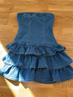 Jeansklänning med volanger och dragkedja - Snygg axelbandslös jeansklänning med volanger i flera lager och markerad midja. Klänningen har dragkedja bak och är i klassisk blå jeans färg. Aldrig använd och säljer då de är alldeles för kort💗 nypris 286 kr