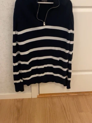 Randig marinblå tröja med dragkedja - Snygg marinblå tröja med vita horisontella ränder och halv dragkedja vid halsen. Tröjan har långa ärmar och ribbade muddar. Perfekt för en avslappnad och stilren look.