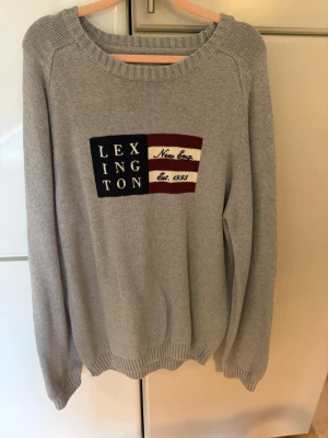 Grå stickad Lexington tröja - Snygg grå stickad tröja från Lexington med broderad logga och flaggdetalj i marinblått, vinrött och vitt på bröstet. Tröjan har rund halsringning, långa ärmar och ribbade muddar. Perfekt för en avslappnad och stilren look.