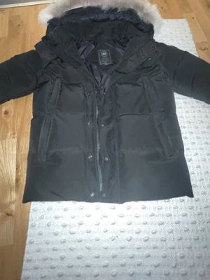Canada goose macmillan - Säljer en svart dunjacka från Canada Goose med stor huva och avtagbar pälskant. Jackan har dragkedja och knappar framtill, två stora fickor och en klassisk puffig design som håller dig varm under kalla dagar.
