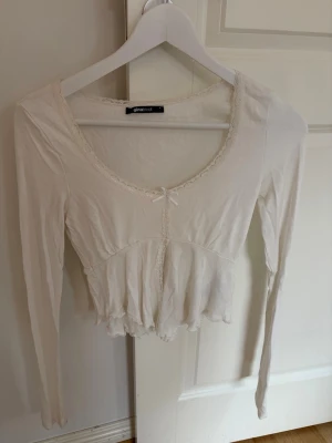 Vit långärmad topp med spetsdetaljer - Söt vit långärmad topp från Ginatricot med spetskant runt ringningen och en liten rosett framtill. Modellen är croppad med lätt volang nedtill och har en feminin, romantisk stil. Perfekt att matcha med höga jeans eller kjol.