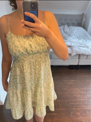 Grön blommig sommarklänning - Söt ljusgrön klänning med vita småblommor och tunna axelband. Klänningen har en rynkad överdel, markerad midja och volangkant nertill. Perfekt för varma dagar och har en luftig, avslappnad passform. Materialet känns lätt och mjukt.