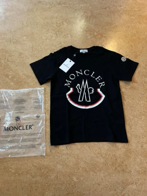 Svart Moncler t-shirt med stor logga - Snygg svart t-shirt från Moncler med stor vit och röd logga framtill. Klassisk rund halsringning och korta ärmar. Tillverkad i mjuk bomull för skön känsla. Helt ny i förpackning. Storlek Small passar någon som har XS