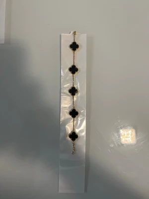 Guldarmband med svarta klöverhängen - Snyggt armband med fem svarta fyrklöverformade hängen infattade i guldfärgad metall. Kedjan är tunn och elegant, och varje hänge har en pärlad kant som ger extra detalj. Perfekt accessoar för att lyfta din outfit.