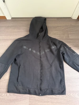 Svart Nike hoodie med dragkedja - Snygg svart hoodie från Nike med hel dragkedja framtill och huva. Diskret Nike-logga på bröstet och ett svart mönster över bröstet. Fickor på sidorna och en extra ficka med dragkedja på ena ärmen. Perfekt för en sportig och avslappnad stil.