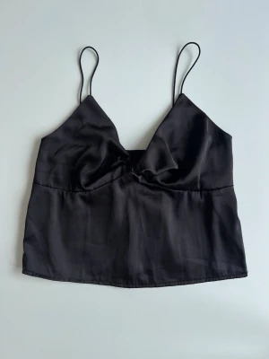 Svart satinlinne med tunna band - nyskick, stl S - En svart clean linnetopp i satin med tunna axelband och fin detalj vid urringningen. Ger en lyxig känsla. Lite croppad i passformen och passar till allt! Knappt använd.