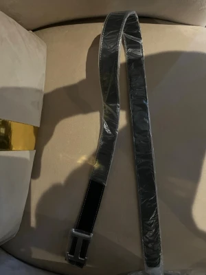 Svart bälte med H spänne från H’er’m”e’s - Snyggt svart bälte från H’er’m”e’s med  H format spänne. Bältet har vita sömmar längs kanterna och är gjort i ett slätt material som ser ut som läder. Perfekt accessoar för att lyfta din outfit.