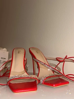 Röda sandaletter med strassdetaljer - Snygga röda sandaletter från Soda med fyrkantig tå, smala remmar dekorerade med glittrande strass och transparenta detaljer. Skorna har höga klackar och tunna snörband som kan lindas runt vristen. Perfekta för att sticka ut och ge en extra touch till outfiten.