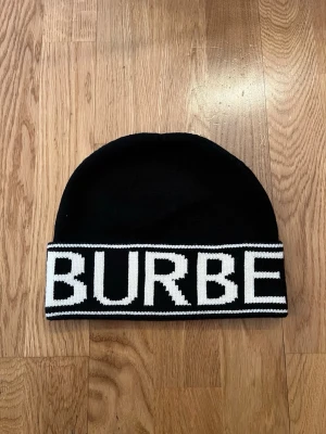 Svart Burberry mössa med logga - Snygg svart mössa från Burberry med bred vit kant där BURBERRY är stickat i stora bokstäver runtom. Klassisk beanie-modell i mjukt stickat material, perfekt för att hålla värmen och ge en trendig look.